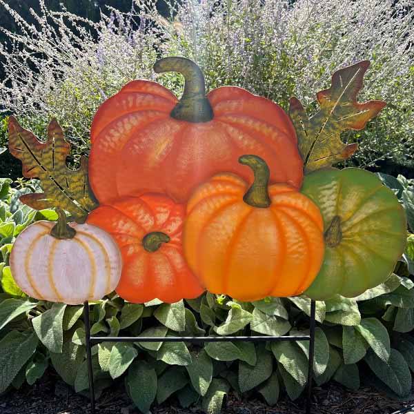 The 28” Pumpkins Stake by Available for Pick-Up & Delivery transforms outdoor spaces into vibrant autumn scenes. The colorful metal pumpkins in orange, green, and white create a lively arrangement when sunlight hits the lush green foliage.