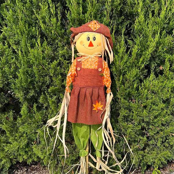 Smiling scarecrow on a bamboo stake wearing an orange patchwork hat and dress with green legs.
