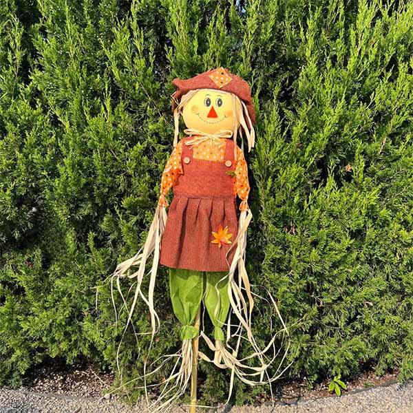 Smiling scarecrow on a bamboo stake wearing an orange patchwork hat and dress with green legs.