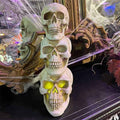 A stack of three 13 Skull Heads with glowing yellow LED eyes from Available for Pick-Up & Delivery sits on a dark surface, with a decorative mirror, cobwebs, and Halloween-themed decor in the background, creating a spooky ambiance.