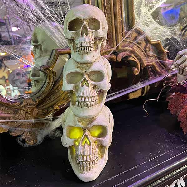 A stack of three 13 Skull Heads with glowing yellow LED eyes from Available for Pick-Up & Delivery sits on a dark surface, with a decorative mirror, cobwebs, and Halloween-themed decor in the background, creating a spooky ambiance.