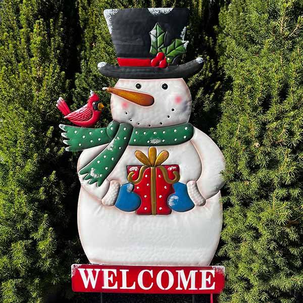 The Snowman Welcome Garden Stake by Available for Pick-Up & Delivery features a cheerful snowman in a top hat and green scarf holding a gift, with a red cardinal perched on its arm. This charming garden stake adds holiday allure, standing among greenery with a WELCOME sign beneath.
