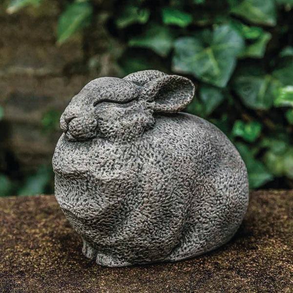 The All Delivery Options Statue – Cast Stone – Bunny features a plump, sleeping rabbit on a brown surface with blurred green ivy in the background, creating a charming garden accent.