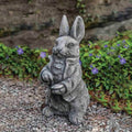 The Statue – Cast Stone – Rabbit Esq. by All Delivery Options features a rabbit in a jacket on gravel, surrounded by purple flowers and greenery—ideal indoor or outdoor décor, with a blurred stone wall in the background.