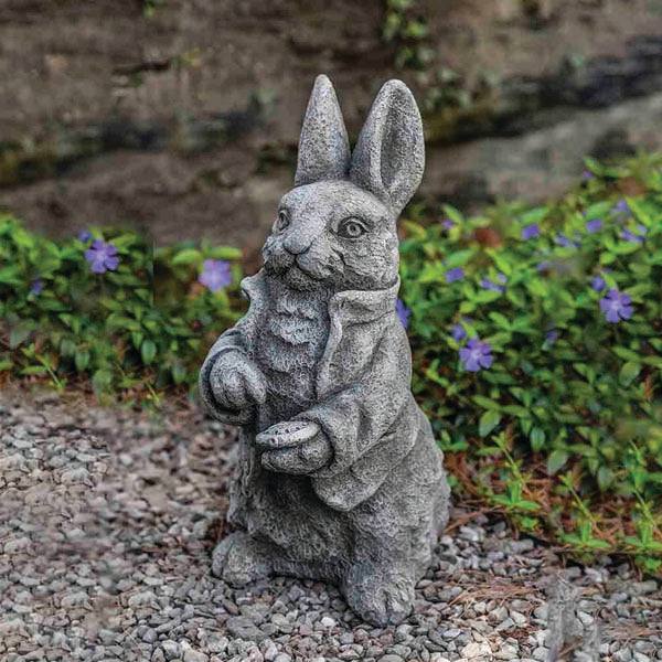 The Statue – Cast Stone – Rabbit Esq. by All Delivery Options features a rabbit in a jacket on gravel, surrounded by purple flowers and greenery—ideal indoor or outdoor décor, with a blurred stone wall in the background.