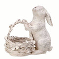 The All Delivery Options Statue – Resin – Bunny with Basket features a white bunny standing on its hind legs, holding a woven basket decorated with vintage-style flowers for rustic charm.