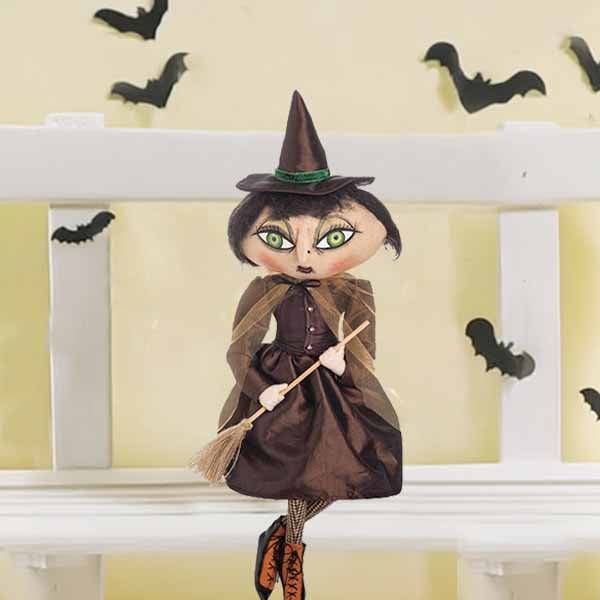 The Joe Spencer whimsical collectible Witch - Matilda - 24 by Available for Pick-Up & Delivery, features a witch with a pointy hat and green eyes on a white bench holding her broomstick. Black bat cutouts flutter against the pale yellow wall, making it an ideal Halloween decoration.